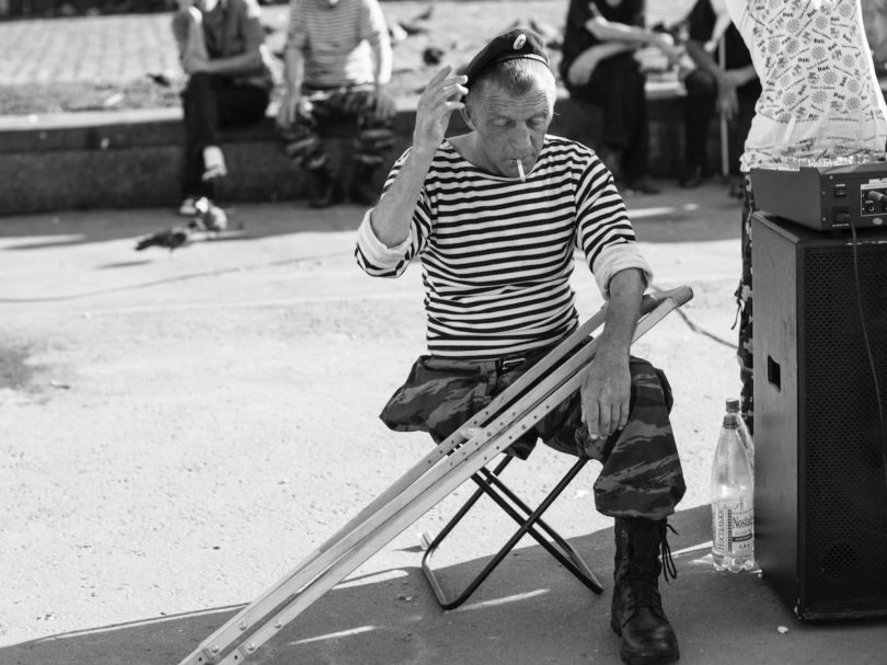 grayscale photo of man sitting on folding stool with crutches in front of people